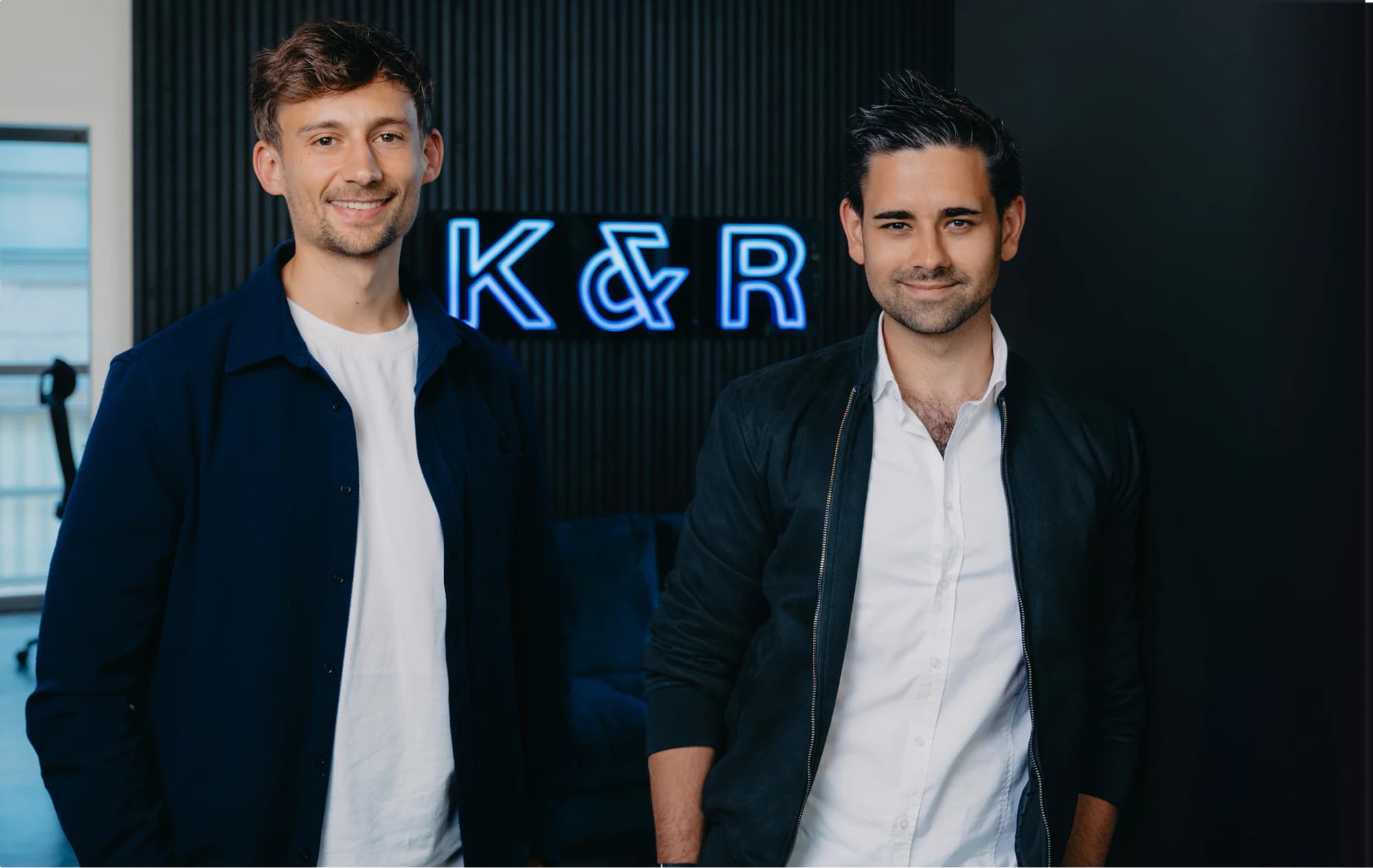 Two men in casual clothes smiling in front of a wall with a glowing blue K&R logo.